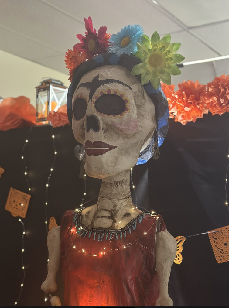 Close-up of a tall Catrina statue with a skeleton face, colorful flower crown, and red dress. String lights and papel picado hang in the background, with orange paper marigolds lining the top edge of the display.