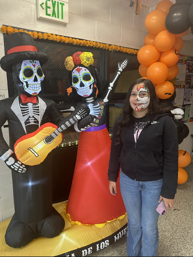 A person with Día de los Muertos face paint stands smiling beside two inflatable skeleton figures dressed as a musician couple. One skeleton wears a suit and top hat and plays a guitar, while the other wears a colorful floral headpiece and long skirt, with one arm raised. Behind them are orange and black balloons and marigold decorations, with an EXIT sign above.