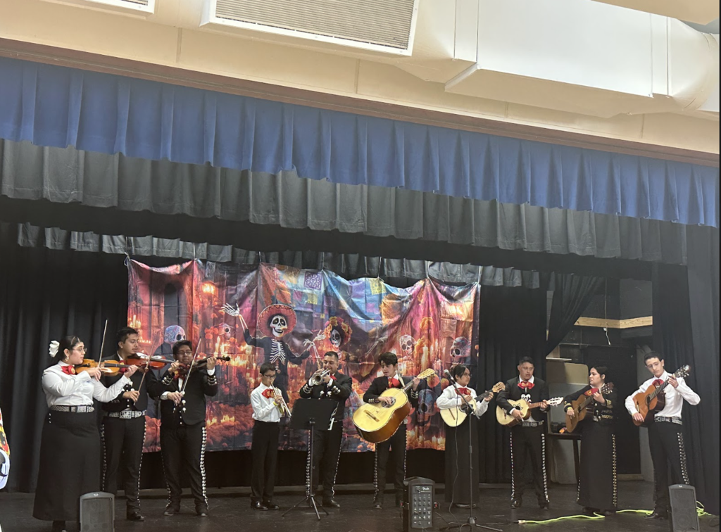 A youth mariachi group performs on a stage, playing violins, trumpets, guitars, and vihuelas. They wear traditional mariachi outfits with red bow ties. The stage backdrop features vibrant Día de los Muertos artwork with skeleton figures and glowing candles.
