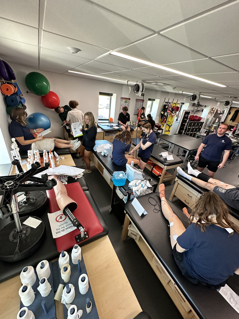 High school students are actively engaged in a sports medicine or athletic training classroom. Multiple students, diverse in gender and appearance, work in small groups at treatment tables practicing taping, stretching, and using equipment like resistance bands and TENS units on each other’s legs. The room contains various medical and athletic supplies including rolls of tape, anatomical models, posters, and large exercise balls mounted on the wall. The environment is bright and hands-on, emphasizing career-based learning in health sciences.