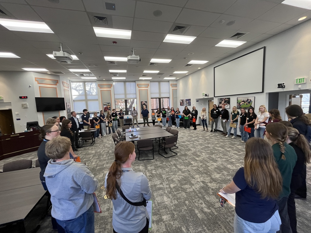 A large group of high school students and a few adults stand in a wide circle inside a spacious boardroom-style meeting room. The students are diverse in appearance and are holding clipboards and folders, suggesting a formal or educational activity. The room features a large projection screen, high ceilings, modern lighting, and wall-mounted displays.