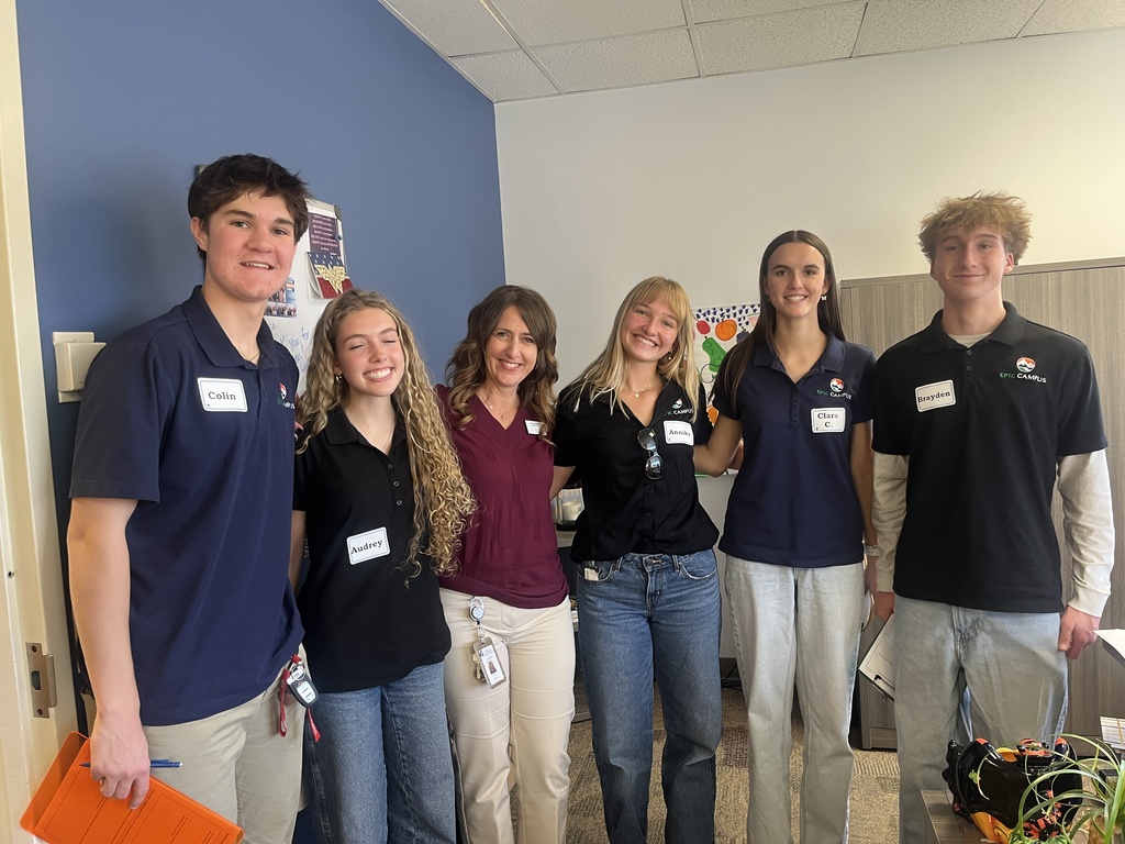 Six people, including four students and two adults, stand smiling for a group photo inside a classroom or office. Each person wears a name tag; the students’ tags read Colin, Audrey, Amilia, Clara C., and Brayden, with some wearing EPIC Campus polos. A poster and colorful artwork are partially visible in the background, along with office furniture. The group appears friendly and engaged.