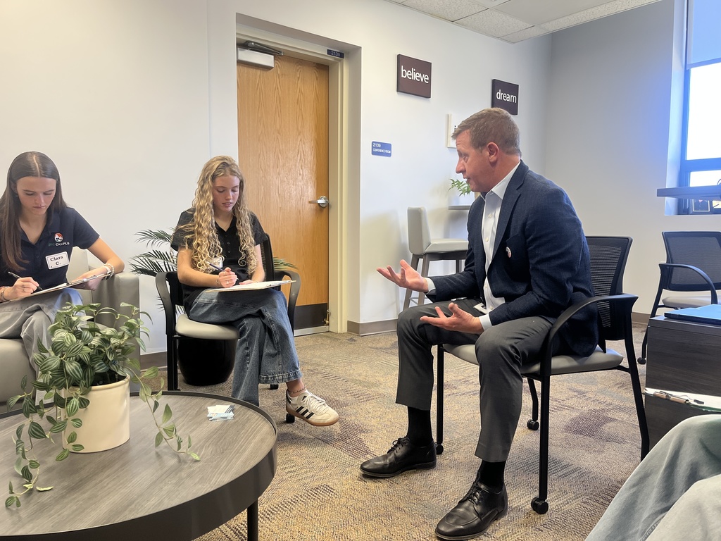 A man in a navy blazer and gray slacks sits in a chair speaking expressively with two high school students, who are attentively taking notes on clipboards. The student on the left wears an EPIC Campus shirt and name tag that reads "Clara C." The room has a modern design with plants and motivational wall signs that say “believe” and “dream.” The setting appears professional and conversational.