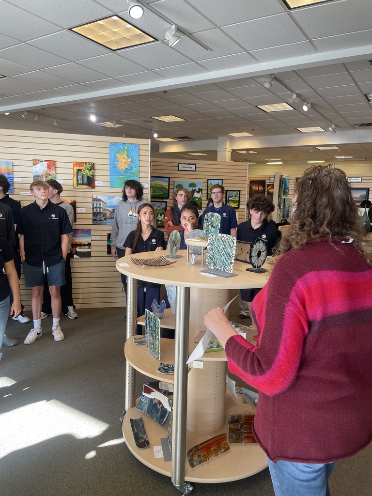 A group of high school students, many wearing black EPIC Campus polo shirts, attentively listen to a woman in a red and pink sweater who is leading a discussion in an art gallery or studio space. The room features bright lighting and walls lined with colorful paintings and artwork. In the center, a circular display shelf holds a variety of fused glass art pieces. The students appear engaged in a field trip or educational tour focused on the arts.