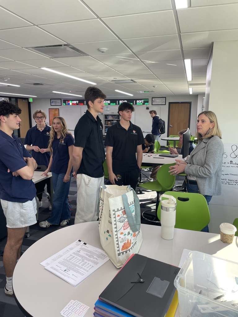 A group of high school students in EPIC Campus polo shirts gather around a female instructor in a modern classroom. They appear engaged in a discussion or receiving instructions. The room features bright lighting, green chairs, and stock market ticker displays above whiteboards, indicating a business or finance-focused setting. On a nearby table are documents, folders, a tote bag, and coffee cups, reinforcing an active, hands-on learning environment.