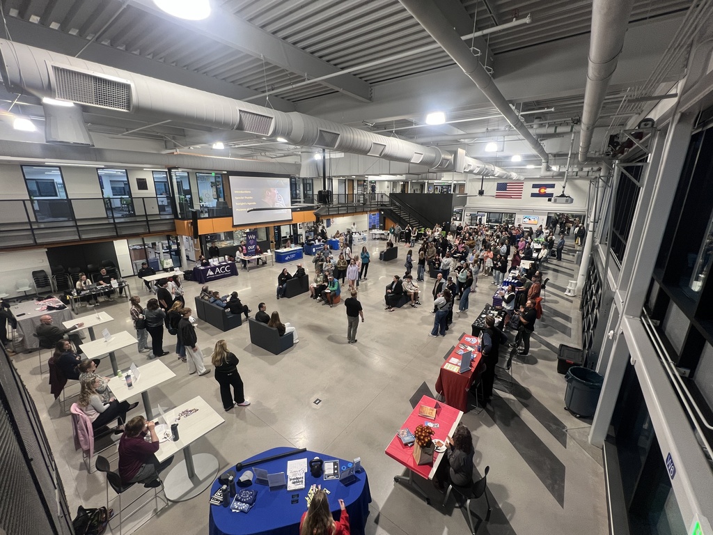 A wide-angle view of a bustling event inside a large, modern school facility with high ceilings and exposed ductwork. The space is filled with tables and booths hosted by various organizations, including Arapahoe Community College (ACC), and is attended by students, families, and staff. A large projector screen displays a presentation near the center, and U.S. and Colorado flags hang on the back wall. Attendees are seated at tables, visiting booths, or standing in groups, indicating a showcase, open house, or college and career fair