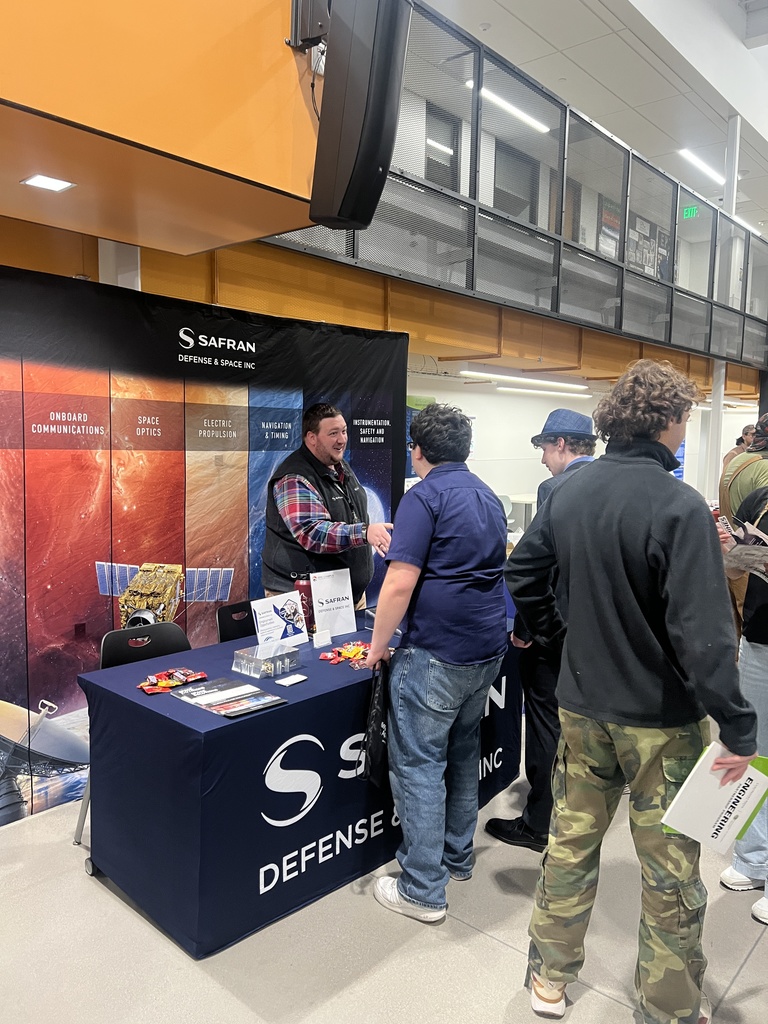 A representative from Safran Defense & Space Inc. engages in conversation with a student at a career fair-style event. The Safran booth features a large, colorful backdrop highlighting their focus areas: Onboard Communications, Space Optics, Electric Propulsion, Navigation & Timing, and Instrumentation, Safety, and Monitoring. The table is covered with a navy blue cloth branded with the Safran logo and has brochures, candy, and promotional items. Several students in casual attire, including one holding a folder labeled “Engineering,” gather around the booth, showing interest in aerospace and defense careers.
