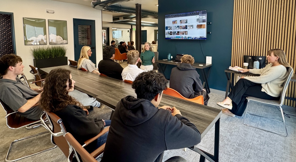 A group of high school students sit around a large conference table in a modern, creative office space, attentively listening to a woman in a green sweater speaking at the front. Another professional, seated to the right, observes while holding papers. A large screen on the wall displays a browser window with search results, possibly part of a presentation. The setting features stylish decor with wood paneling, plants, and contemporary furnishings, suggesting a professional development or workplace learning experience.