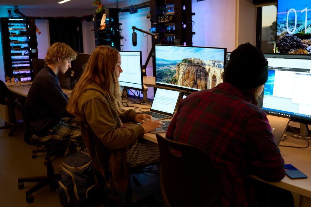 Three high school students work independently at a long desk in a darkened, tech-focused classroom. Each student uses multiple monitors and laptops displaying coding interfaces, web content, and desktop environments. The room includes illuminated server racks, overhead microphones, and ambient blue and purple lighting, suggesting a cybersecurity or IT learning lab. The students appear focused and engaged with their tasks.