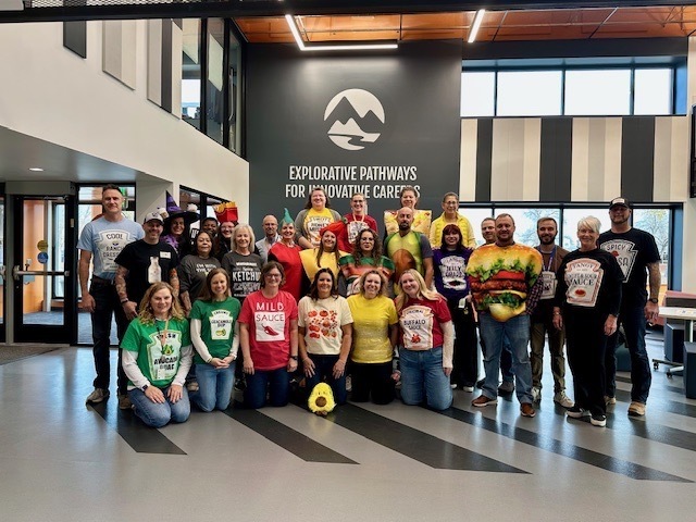 A large group of EPIC Campus staff pose together in the school’s lobby, standing in front of a wall with the EPIC logo and the words "Explorative Pathways for Innovative Careers." Everyone is dressed in fun food-themed costumes or shirts, including items like ketchup, mustard, mild sauce, guacamole, burgers, hot dogs, and other condiments. The group is smiling and festive, celebrating a themed spirit day in a modern, brightly lit school environment.
