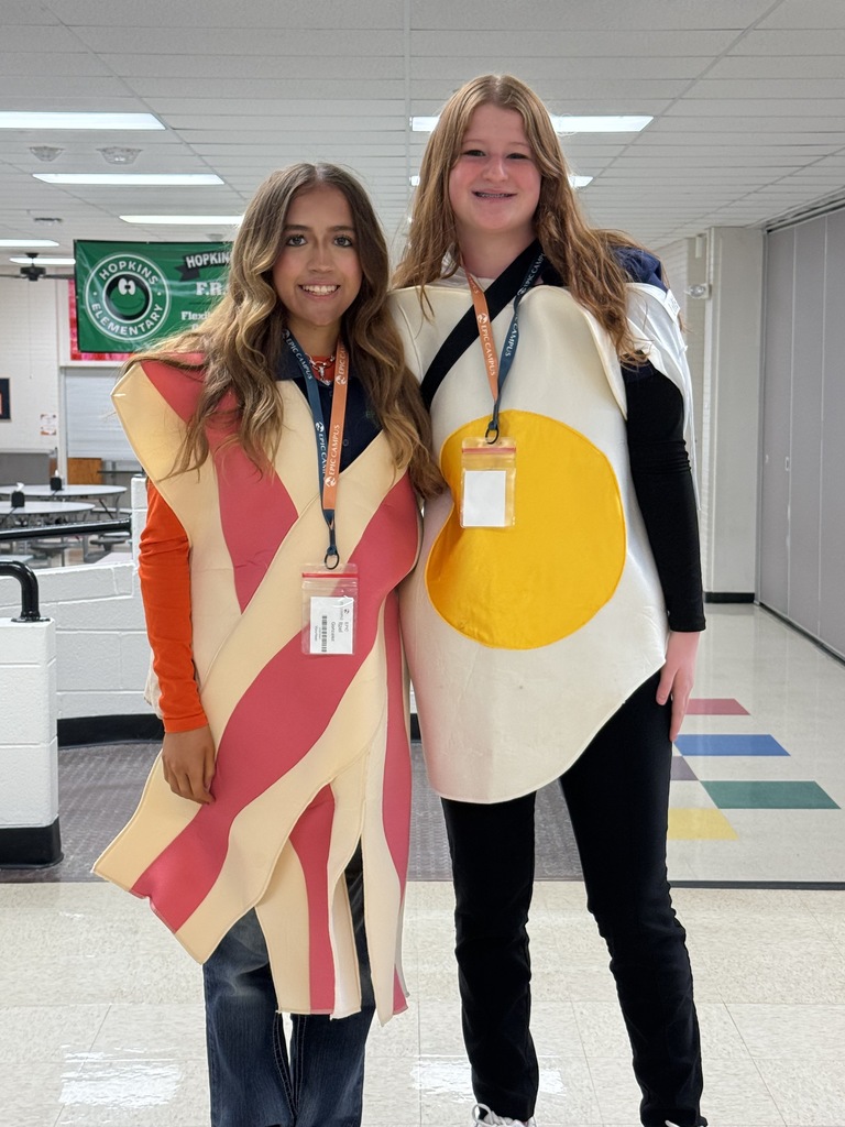 Two high school students stand smiling in a school hallway, wearing themed costumes for spirit day. One is dressed as a strip of bacon and the other as a sunny-side-up egg. Both wear EPIC Campus lanyards with ID badges. The background shows part of a cafeteria area with white brick walls, colorful floor tiles, and a green “Hopkins Elementary” banner hanging from the ceiling.