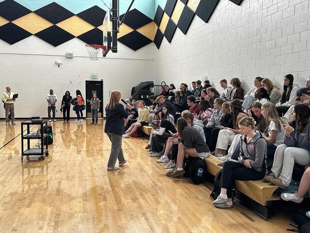 Students seated on bleachers in a gym, listening to a presentation by an adult woman walking in front of them.