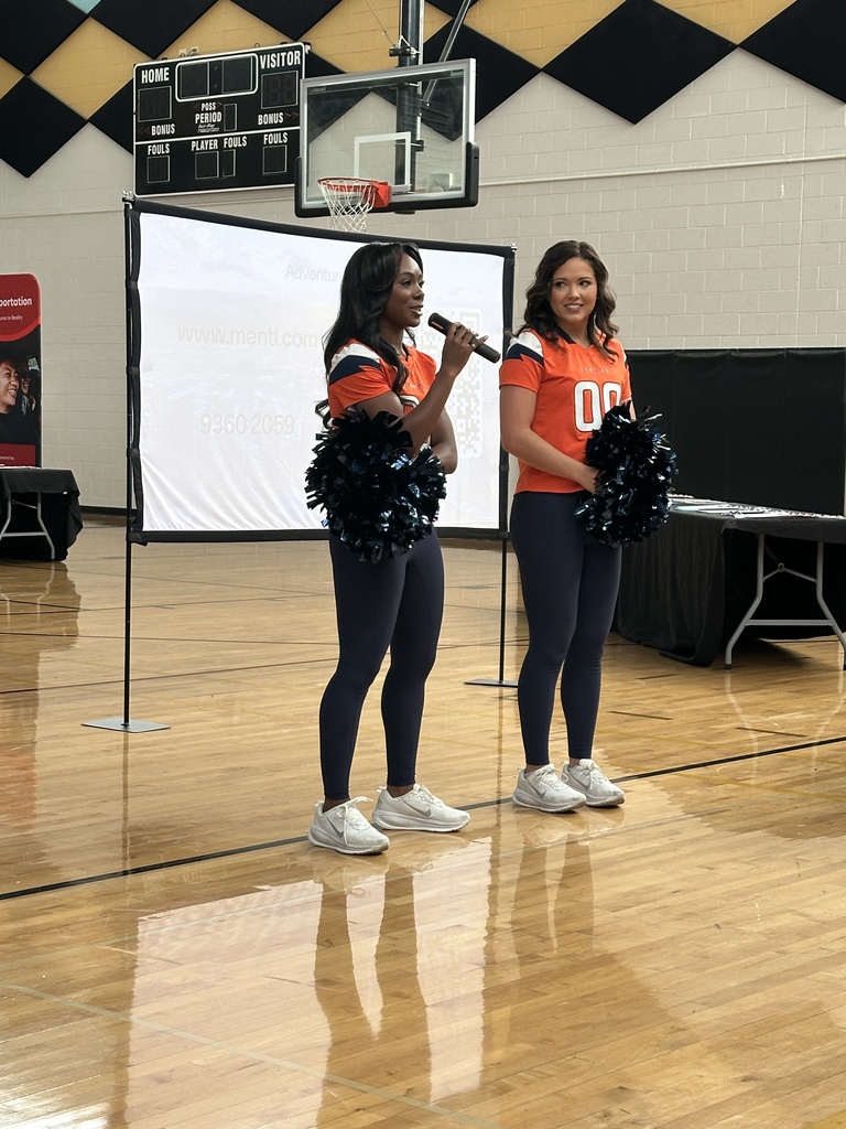 Two women in Denver Broncos cheerleading outfits stand in front of a screen on a gym floor, one holding a microphone.