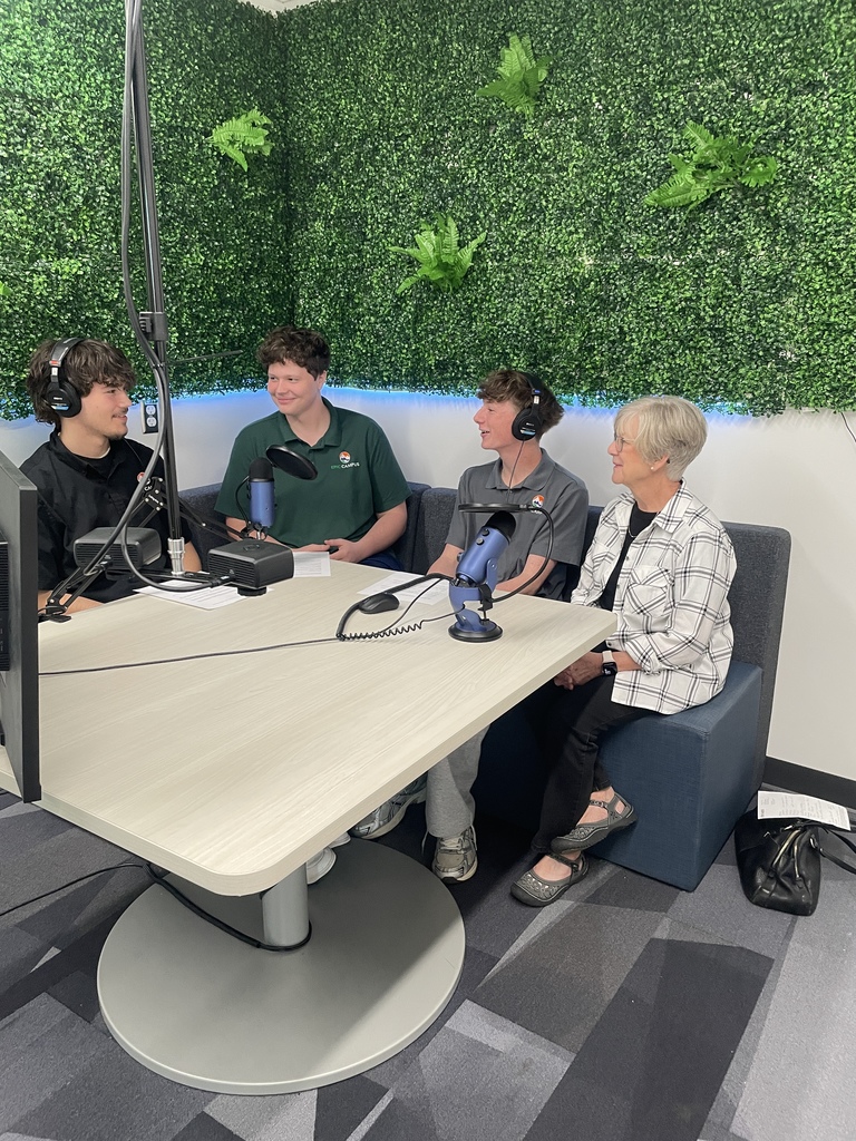Four people—three students and an older woman—sit around a table in a small recording studio with a faux greenery wall and mounted ferns. All three students wear headphones and speak into professional podcast microphones. Two students wear Littleton Public Schools shirts with visible LPS apple logos. The older woman, dressed in a plaid shirt and black pants, sits on a blue bench next to them. The space is brightly lit, with a modern carpet and recording equipment on the table.