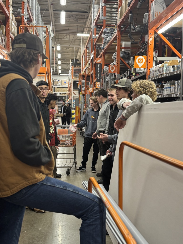 A group of high school students stands in an aisle of a Home Depot, gathered around a flat cart loaded with large drywall sheets and metal pipes. The students appear to be on a field trip or hands-on learning activity, engaged in conversation. Some wear sweatshirts with school or program logos, including one that reads “Heritage Construction.” The store's tall orange shelving units are filled with boxes, electrical supplies, and tools. Overhead signs mark the aisles, with visible signs for “Electrical Fittings” and aisle numbers 9 and 10. The environment is bright with overhead lighting.