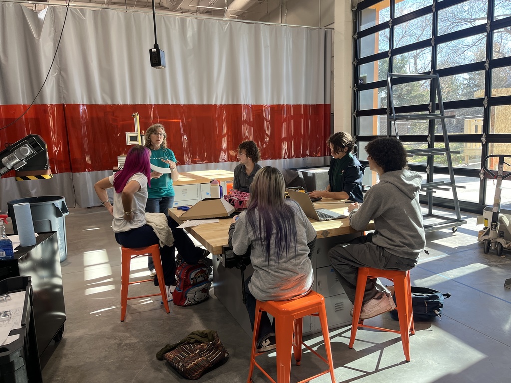 A group of six high school students, diverse in gender and appearance, sit on bright orange stools around a workbench in a sunlit makerspace or tech lab. An instructor stands at the front, gesturing while speaking to the group. Some students have laptops open, and materials like cardboard, tools, and water bottles are on the table. The room features a red and white industrial curtain, tall windows with natural light streaming in, a metal ladder, and garage-style doors. The space suggests hands-on, project-based learning.