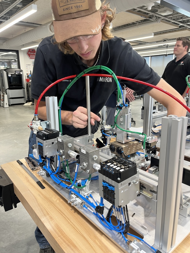 A student wearing safety glasses and a brown cap labeled “J.B. Holmes Since 1961” works intently on a pneumatic automation system in a technical classroom or lab. The student is using tools to adjust the machinery, which is composed of metal components, wiring, and color-coded air tubes (red, green, and blue). His dark shirt has a visible “MIKRON” logo and a name tag that reads “Holden.” Another person in the background, also in dark clothing with an LPS (Littleton Public Schools) logo, observes the activity. The space is modern, with industrial lighting and equipment throughout.