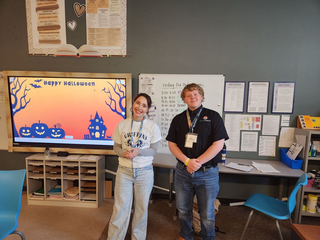A teacher and a student stand smiling in front of a classroom screen with a Halloween-themed slide reading “Happy Halloween.” The teacher wears a “GRIFFINS” T-shirt, and the student wears a black LPS Career Campus shirt with a name badge. Behind them are whiteboards filled with schedules and instructional posters.