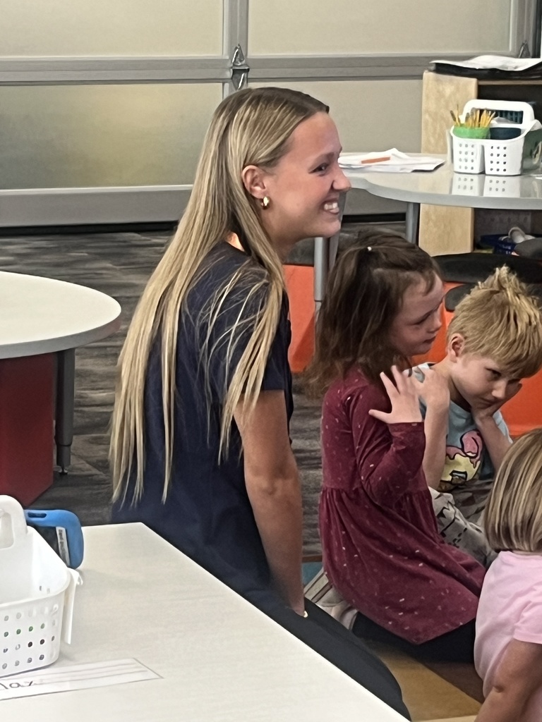 A young woman with long blonde hair, wearing a navy shirt, kneels beside a small group of young children in a brightly lit classroom. She smiles warmly while the children—two girls and a boy—sit on the floor, engaged and interacting. The classroom has white tables, orange seating, and organized bins of supplies.