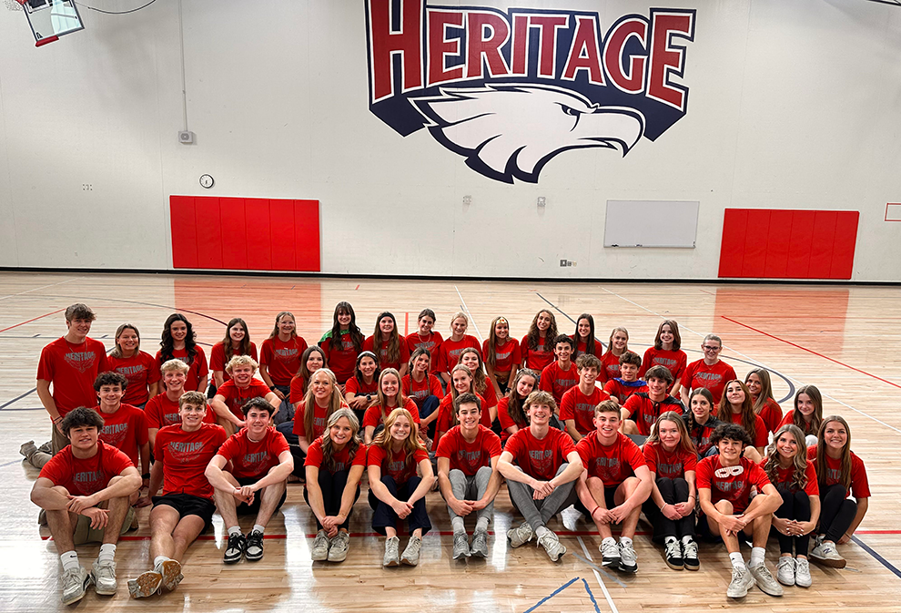 Large group of high school students wearing matching red “Heritage” shirts pose together in a gymnasium. Most are seated cross-legged on the polished gym floor, with a few standing in the back. A large “HERITAGE” wordmark with an eagle mascot is painted on the wall behind them in red, white, and blue. Red wall mats, a whiteboard, and a basketball hoop are visible in the background.
