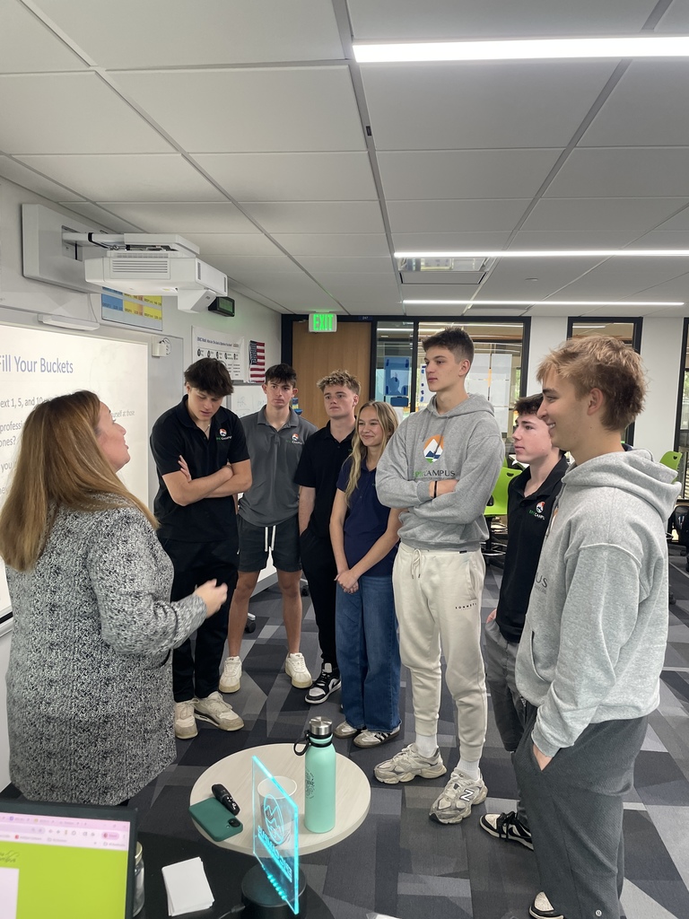 A group of seven high school students, diverse in gender and appearance, stand in a semicircle facing a woman who appears to be a teacher or presenter. They are inside a modern classroom with white walls, gray carpet tiles, and fluorescent lighting. The teacher is speaking near a projection screen that displays a partially visible slide titled “Fill Your Buckets.” Several students wear shirts or hoodies with the "EICampus" logo. A small round table in the foreground holds a teal water bottle and an acrylic award. The atmosphere appears attentive and engaged.