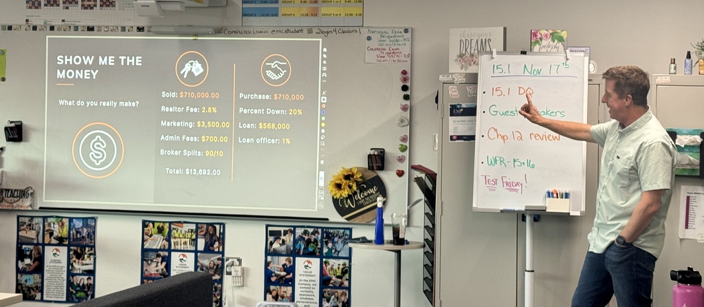 A male speaker stands near a whiteboard and points toward it while explaining a projected slide titled “SHOW ME THE MONEY.” The slide outlines earnings from a real estate transaction, with figures including $710,000 sold and purchased, and a $13,692 total income after fees. The whiteboard to the right includes the date “Nov 17th” and class topics like “Chp 12 review” and “Test Friday!” The room features posters, a sunflower decoration, and student-focused displays along the walls.