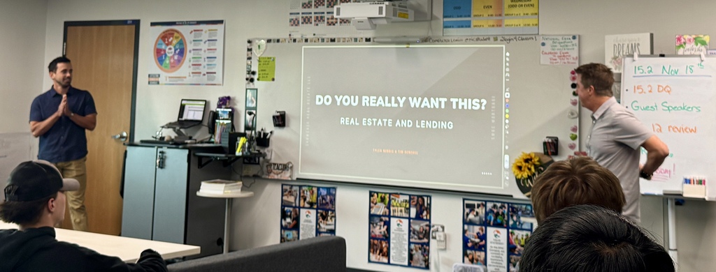 A classroom presentation is in progress with two adult male guest speakers and several students watching. The slide on the projector reads “DO YOU REALLY WANT THIS? REAL ESTATE AND LENDING.” One speaker stands to the right of the screen mid-sentence, while the other stands by the door, hands clasped. A whiteboard to the right lists the date “Nov 18th” and topics such as “Guest Speakers” and “Chp 12 review.” Classroom walls display colorful posters, student work, and educational materials.