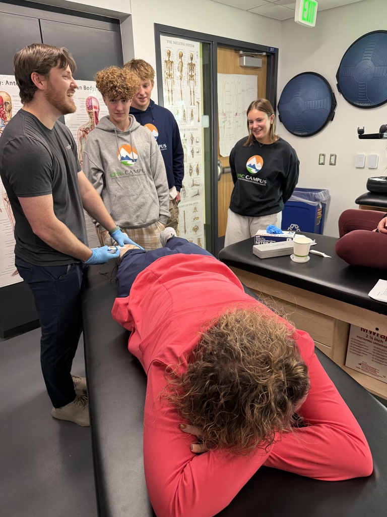 In a classroom decorated with anatomical posters, a man in gloves demonstrates a physical therapy or athletic training technique on a person lying face-down on a table. Three students in EPIC Campus hoodies and sweatshirts observe closely, smiling and engaged. The person on the table wears a red sweatshirt and has curly hair. The space includes therapy equipment like BOSU balls on the wall, and medical supplies on a side counter.