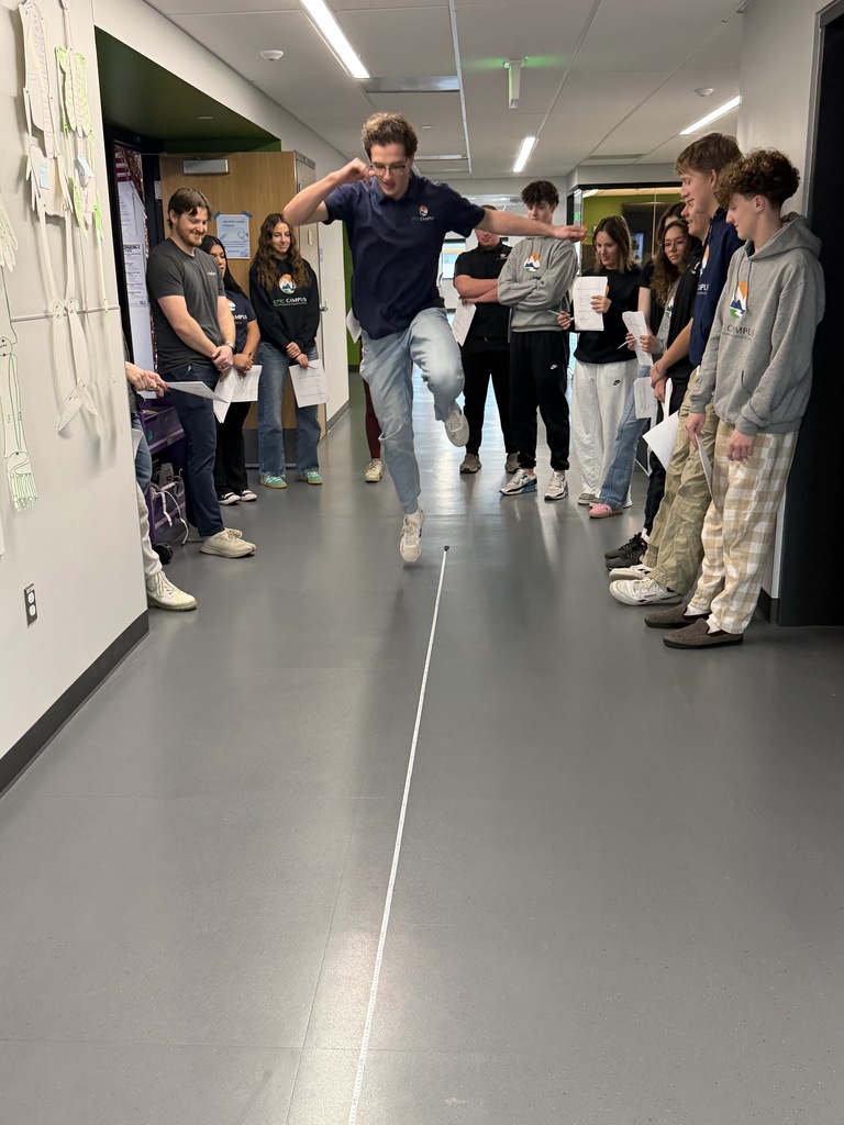 A student is mid-jump in a hallway activity, leaping over a long measuring tape stretched on the floor. He is wearing a navy EPIC Campus shirt and jeans, with arms raised for balance. Students and an adult stand along both walls watching, some holding papers. The group is diverse in gender and appearance, with most wearing EPIC Campus apparel. Drawings of bones and anatomical illustrations are visible on the wall to the left, suggesting a health or science context.