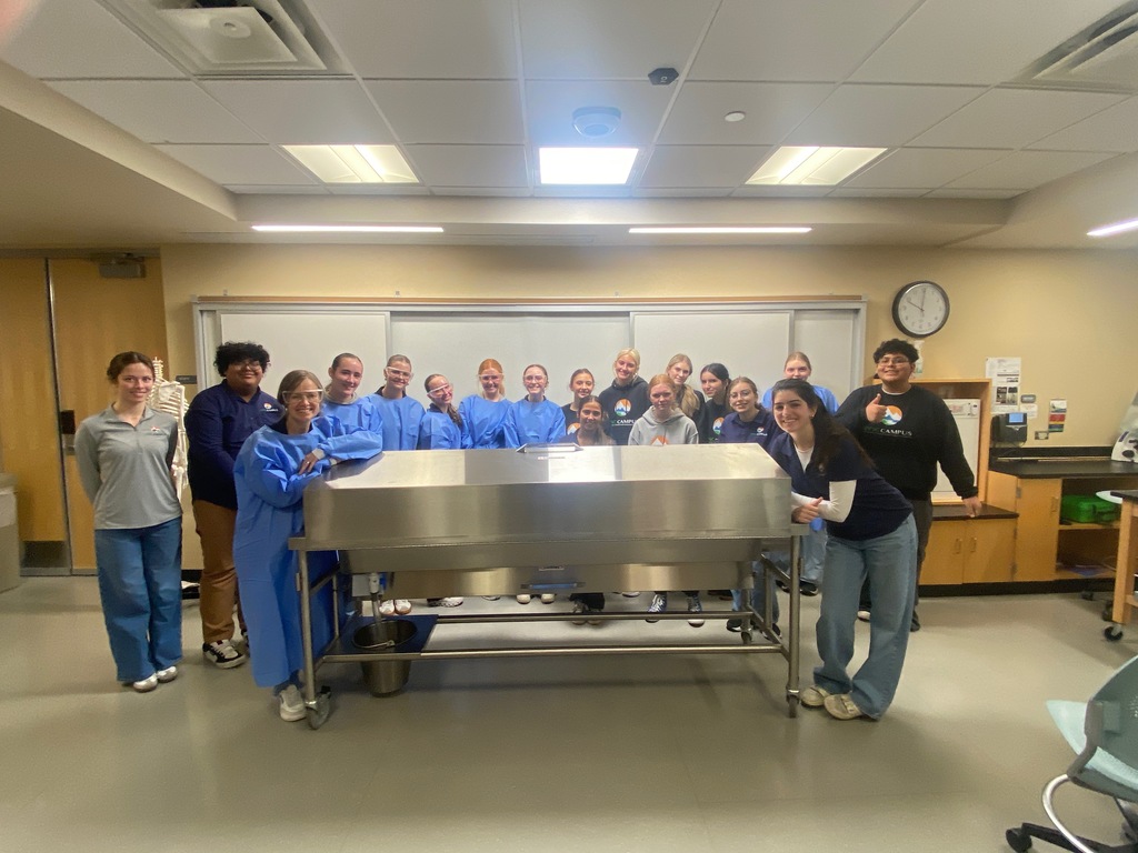 A group of students and educators pose around a stainless steel cadaver table in a classroom. Most individuals wear blue surgical gowns, while others wear casual or branded school attire with logos showing “EPIC CAMPUS.” The background features lab furnishings, a whiteboard, and a clock. One student gives a thumbs-up gesture.