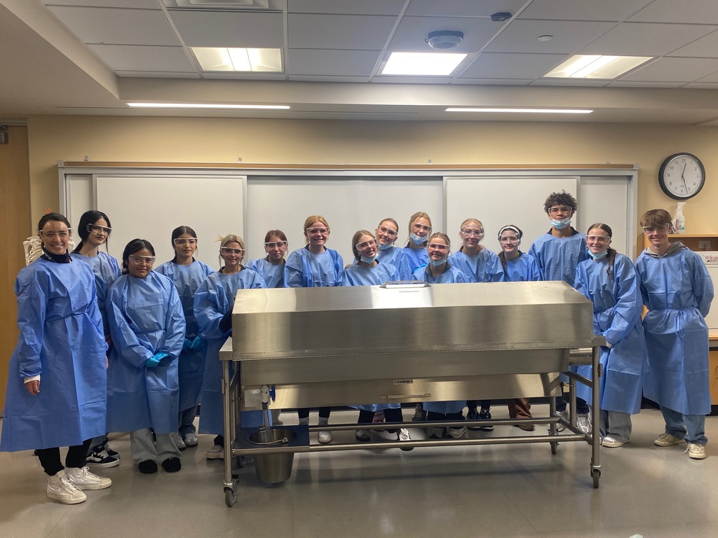 Seventeen high school students stand around a metal cadaver table in a classroom, all dressed in blue surgical gowns and wearing protective eyewear. A few students have face masks under their chins. The room has beige walls, whiteboards, a clock, and lab equipment in the background.