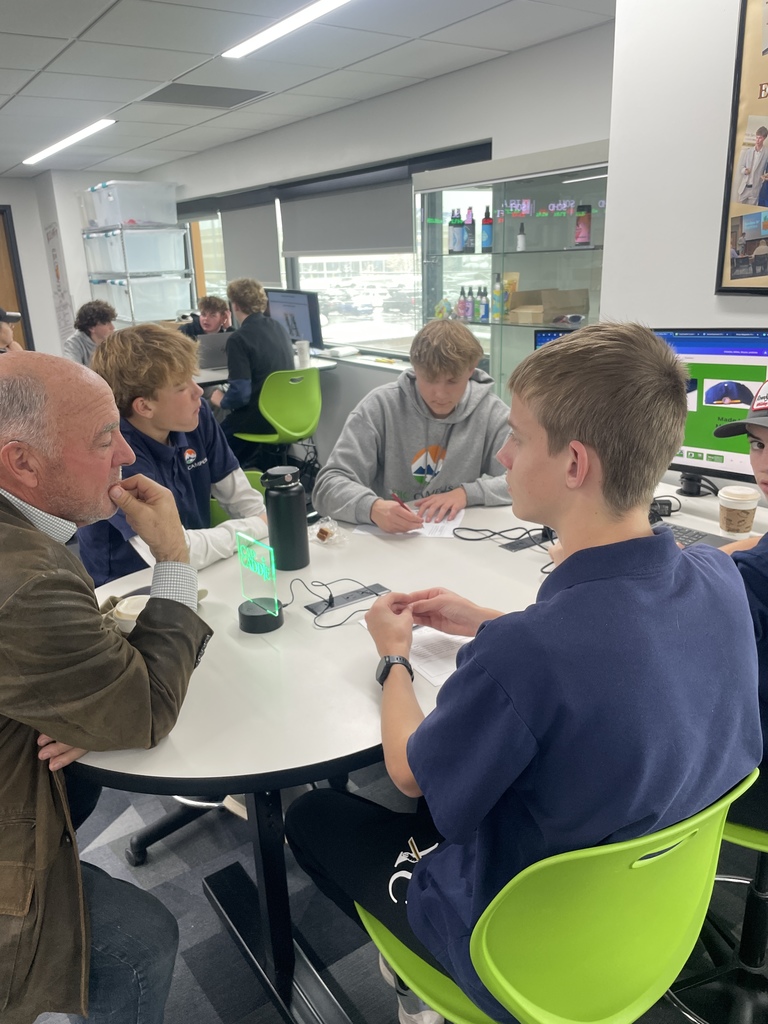 An older adult man in a brown blazer sits with four high school students at a round table, all actively engaged in discussion. Some students wear “EPIC Campus” gear. The table has earbuds and devices for interviews or audio work. Behind them are large windows, a display case with student products, and more students working on laptops.