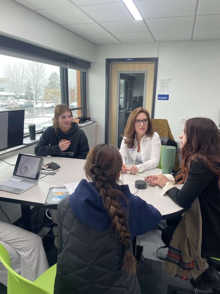 Four students and one adult sit around a round table in a bright classroom with large windows showing snowy trees outside. The group appears engaged in discussion. One student wears an “EPIC Campus” hoodie, and laptops, notebooks, and water bottles are on the table. A door labeled "247A" and a sign reading “Recording” are visible in the background.