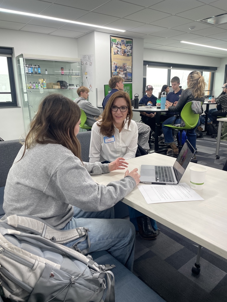 An adult woman wearing glasses and a name tag labeled "Jana" sits at a low table across from a student with long hair and a backpack. A laptop and printed worksheet are on the table. Other students work in the background at tables with green chairs. A display cabinet of student products and an “Entrepreneurship” poster are visible on the wall.