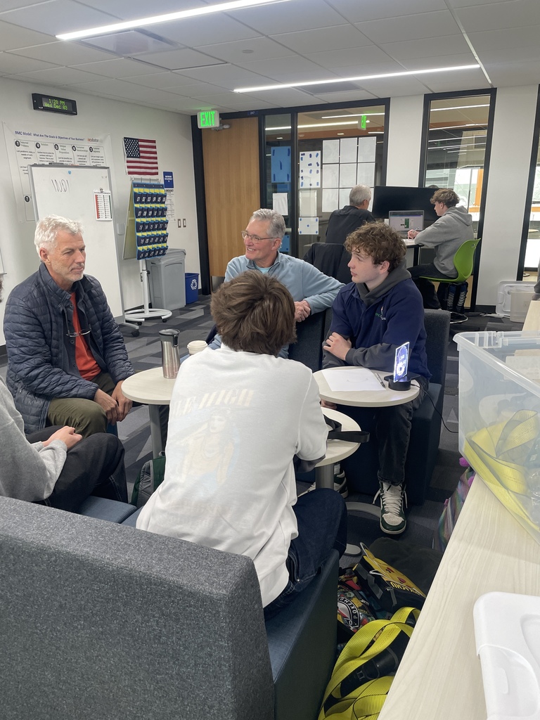 Two adult men and three high school students sit in a group at small round tables in a collaborative classroom space. The group appears to be in conversation. The room has modern furniture, visible signage related to business education, and an American flag. Students in the background work on computers at additional stations.