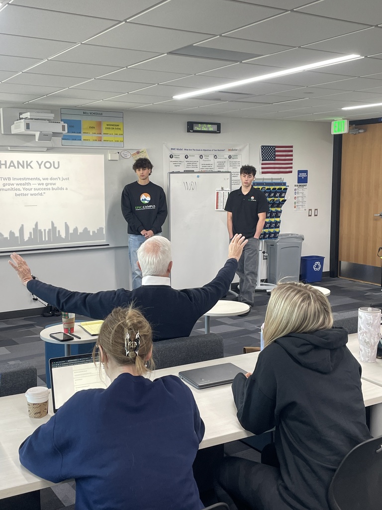 Two high school students stand at the front of a classroom giving a presentation, with a slide on the screen that reads "THANK YOU" and includes a message about growing communities. One wears an EPIC Campus sweatshirt, the other a polo with the same logo. An adult in the audience raises both arms in encouragement, while others watch from behind laptops and notebooks. The room features educational posters, a whiteboard, and an American flag.