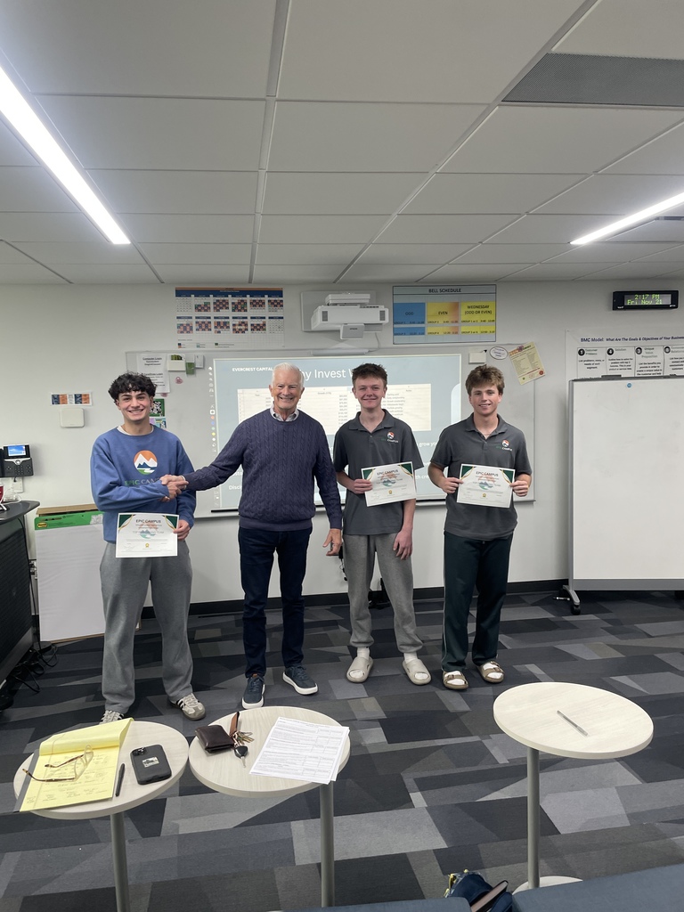 Three students and one adult stand at the front of a classroom smiling for a photo. Each student holds a certificate labeled “EPIC Campus.” Behind them is a presentation slide titled “Evercrest Capital” and a projector mounted on the ceiling. The adult shakes hands with one of the students in congratulations. Classroom furniture and presentation materials are visible in the foreground.
