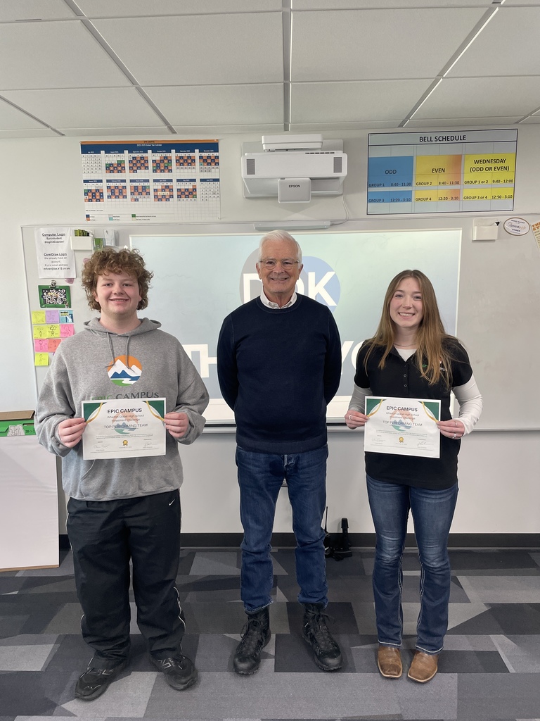 A male and female student stand on either side of an adult, all smiling and holding certificates labeled “EPIC Campus – Top Performing Team.” Behind them is a projection screen showing the EPIC Campus logo, along with a wall-mounted projector, colorful school schedules, and posters. The setting is a classroom with a professional, celebratory atmosphere.
