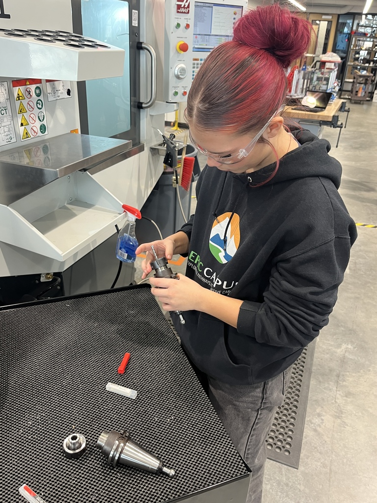 A student with vibrant red hair tied up in a bun works with metal machine components at a black work surface inside a machining lab. She wears protective goggles and an “EPIC Campus” hoodie. A CNC machine and precision tools are visible nearby, indicating a manufacturing or engineering-focused setting
