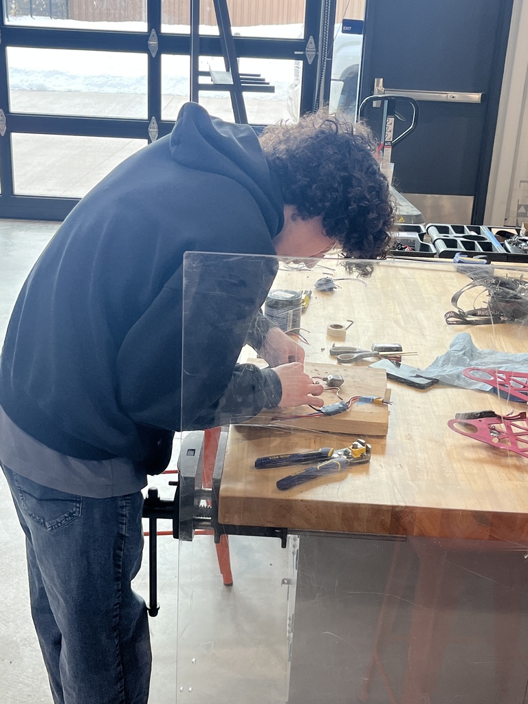 A student wearing a dark hoodie and jeans leans over a wooden workbench, focused on assembling or wiring a small device. Various tools, wires, and components are spread out on the bench. The space has large garage-style windows showing snow outside and is equipped with workshop tools and safety features, including a clear plastic safety shield.