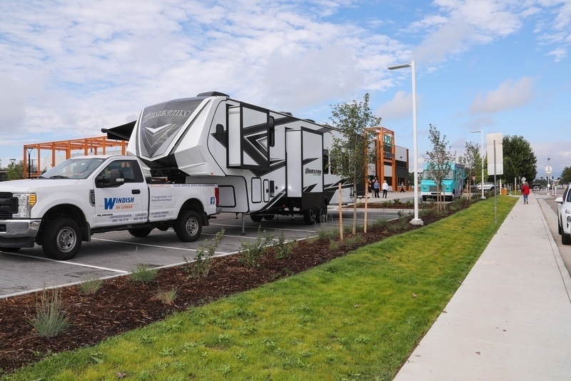 A fifth-wheel Momentum RV is hitched to a Windish RV pickup truck in a parking lot with grass and modern buildings in the background.