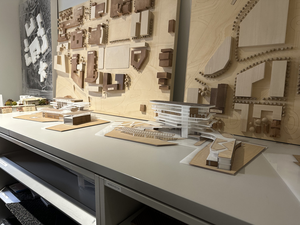 A collection of architectural models made of wood and acrylic are arranged on a white shelf. Behind the models are large, detailed site maps and building layouts mounted on the wall, showcasing urban and campus planning. The models include multi-story buildings and surrounding landscape features, emphasizing architectural design and spatial planning.