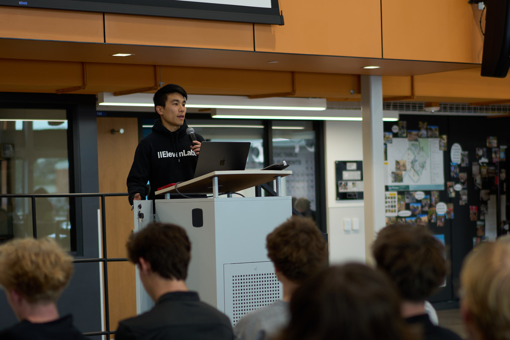A young male speaker stands at a podium, holding a microphone and speaking to an audience of students. He wears a black hoodie with the text “IIElevenLabs” on it. Behind him is a laptop and a modern school interior with orange accents and wall displays. Students' heads are visible in the foreground as they listen.