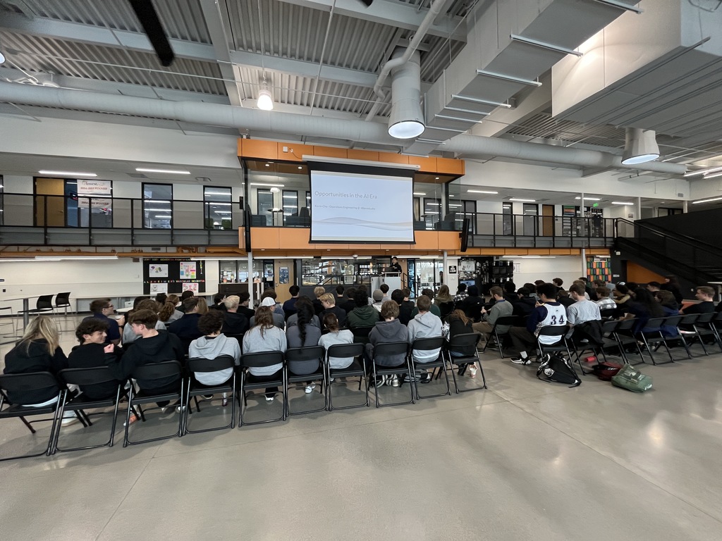 A large group of high school students sit on folding chairs in a spacious, modern school atrium with high ceilings and exposed ducts. A presentation titled “Opportunities in the AI Era” is projected on a screen above a podium. The atrium features two levels with glass-walled rooms and a staircase to the right, with posters and student work displayed on the lower wall.