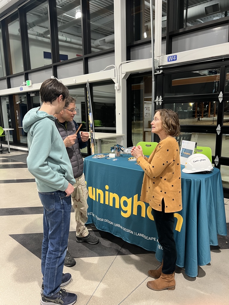 Two students speak with a woman at a booth representing Cunningham, an architecture and design firm. The table is covered in a teal cloth with the firm’s logo and includes pamphlets, giveaways, and a white hard hat. They are in a brightly lit school space with large windows and green chairs visible in the background.