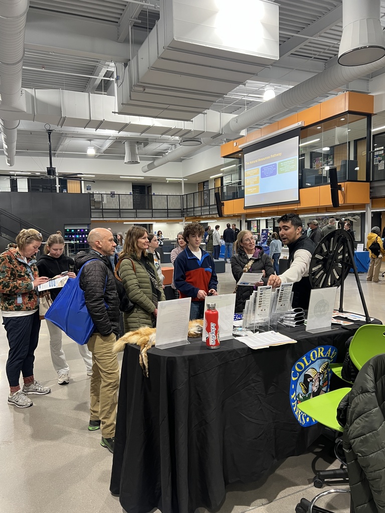 A representative from Colorado Parks and Wildlife speaks with attendees at a booth draped in a black tablecloth with the agency’s seal. Displayed materials include brochures, animal pelts, and educational items. Several students and adults stand in line, engaging with the representative beneath a projection screen about career pathways.