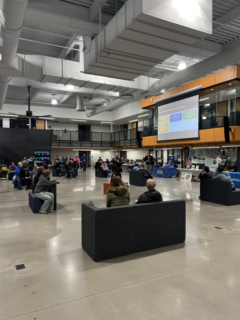 Students, families, and educators are gathered in a modern school atrium for a career pathways event. People are seated on modular couches and chairs, facing a large screen that displays a presentation titled “The Natural Resources Pathway.” Multiple booths and tables are set up around the space, creating a collaborative and informative environment.