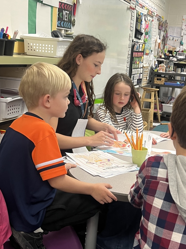 A close-up of the same group from Image 1 shows a student leader helping younger children review colorful worksheets, likely part of a reading or art activity. The classroom is vibrant, with a whiteboard, bins, and posters in the background. The interaction is warm and focused.