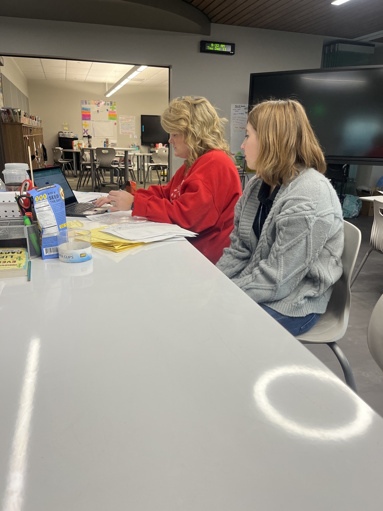 An adult teacher and a high school student sit side by side at a white table in a modern classroom. The teacher is helping the student with paperwork, both focused on the task. Supplies like sticky notes, a laptop, and snacks are on the table. In the background, another classroom space with posters and a large screen is visible.
