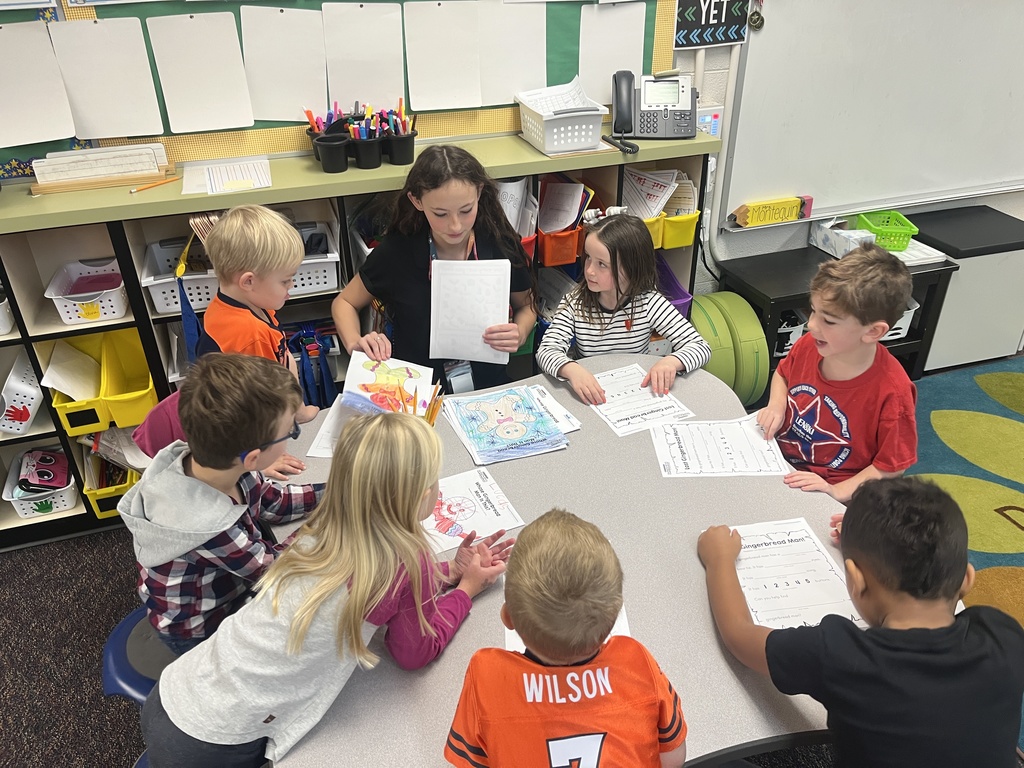A group of young elementary students sits around a circular table with a student leader, likely an older peer, guiding an activity. The table is covered with colorful worksheets and art. The students are engaged and focused. Shelves with bins, supplies, and educational materials line the background in a bright, welcoming classroom.